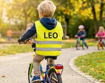 Puede incluir: Un niño con un chaleco de seguridad amarillo con el nombre "LEO" impreso monta en bicicleta en un camino pavimentado. El chaleco tiene rayas reflectantes. Otros niños montan en bicicleta en el fondo.