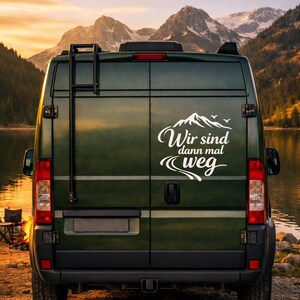 May include: A dark green camper van featuring a white decal on the rear doors. The decal displays a mountain graphic, birds, and the German text "Wir sind dann mal weg" with a winding road illustration. The van includes a black rear ladder.