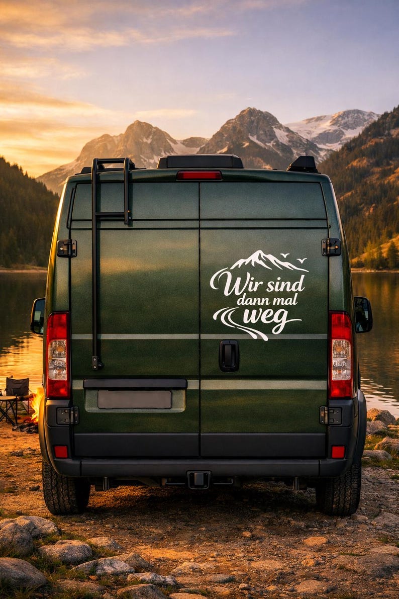 May include: A dark green camper van featuring a white decal on the rear doors. The decal displays a mountain graphic, birds, and the German text "Wir sind dann mal weg" with a winding road illustration. The van includes a black rear ladder.