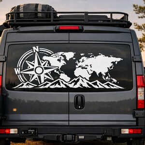 Puede incluir: Una furgoneta camper de color gris oscuro con un gran vinilo blanco en la luna trasera. El dise&ntilde;o muestra un mapa del mundo detallado, una rosa de los vientos con las letras N y W, y una silueta de monta&ntilde;as en la parte inferior del cristal.