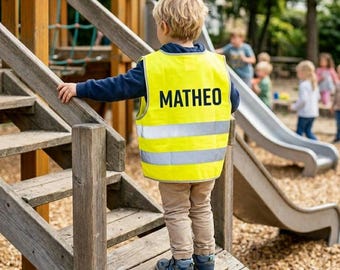 Puede incluir: Un niño lleva un chaleco de seguridad amarillo brillante con el nombre "MATHEO" impreso en negro. El chaleco tiene dos franjas plateadas reflectantes. El niño está en una estructura de juego de madera.