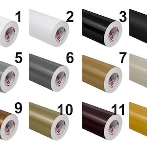 May include: A grid of twelve rolls of adhesive vinyl film, each labeled with a number from 1 to 12. The rolls display a variety of solid colors including white, black, gray, beige, brown, gold, and yellow, all featuring a white inner core.