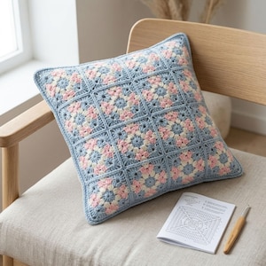 May include: A square crochet pillow cover constructed from multiple granny squares in shades of light blue, soft pink, and cream. The pillow is accompanied by a printed crochet pattern instruction booklet and a wooden handled crochet hook.