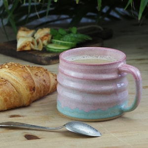 Könnte beinhalten: Handgefertigte Keramiktasse mit rosa und türkis Glasur, gefüllt mit einem hellen Getränk. Ein Croissant, Gebäck und ein Löffel liegen auf einer Holzoberfläche, was auf ein Frühstück hindeutet. Die Tasse hat ein strukturiertes, geriffeltes Design.