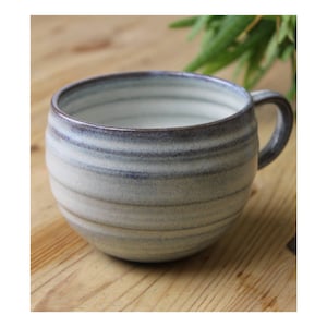 Può includere: Tazza in ceramica fatta a mano con forma arrotondata e manico. La tazza presenta una combinazione di colori azzurro chiaro e crema con linee orizzontali strutturate. Il bordo e il manico sono blu scuro. La tazza è progettata per bevande calde.