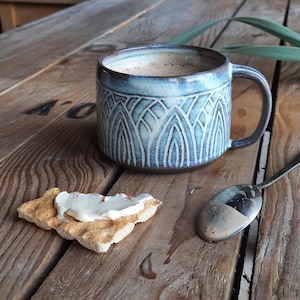 Può includere: Una tazza in ceramica con un motivo geometrico blu e bianco, piena di una bevanda calda. Un pezzo di pane con formaggio cremoso e un cucchiaio con residui di caffè sono su una superficie di legno.