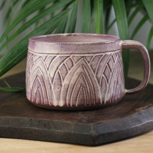 Könnte beinhalten: Handgefertigte Keramiktasse in Mauve- und Cremetönen mit einem geometrischen Art-Deco-Muster. Die Tasse hat einen Henkel und steht auf einer dunklen Holzoberfläche, mit grünen Blättern im Hintergrund. Ein Unikat für Kaffee oder Tee.