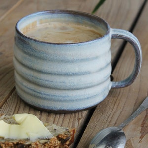 Op de afbeelding: Een lichtblauwe keramische mok met een geribbeld ontwerp, gevuld met koffie. Een sneetje brood met boter en een lepel liggen op een houten oppervlak. De mok heeft een bruine rand en een handvat.