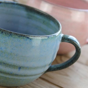 Op de afbeelding: Close-up van een handgemaakte keramische mok met een blauwgroen glazuur en een gebogen handvat. De mok heeft een getextureerd oppervlak en staat tegen een wazige achtergrond met een andere roze mok. Het aardewerk staat op een houten oppervlak.
