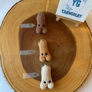 May include: Three crocheted dog bone keychains in different colours: brown, tan, and white. Each keychain has a black button nose and two black eyes. The keychains are displayed on a wooden surface with a small sign that says "YG YARNCOLBY".