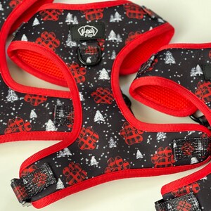 May include: Three red and black dog harnesses with a paw print and Christmas tree pattern. The harnesses have a red adjustable strap and a black buckle. The harnesses are labeled "Woof Pup".