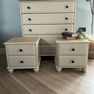 Op de afbeelding: Een crèmekleurige slaapkamerset bestaande uit een hoge ladekast en twee bijpassende nachtkastjes. De meubels hebben zwarte knoppen en lichte houten bladen. Elk nachtkastje heeft twee lades. De set staat op een houten vloer.