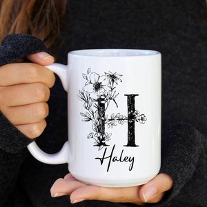 Peut inclure: Une tasse en céramique blanche avec un motif floral noir et la lettre "H" entourée de fleurs. Le nom "Haley" est écrit sous le motif.