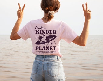 Puede incluir: Una mujer con una camiseta rosa con el texto "Create a Kinder Planet" y un gráfico de una Tierra en forma de corazón con un anillo alrededor. El texto debajo del gráfico dice "The world could always use more kindness."