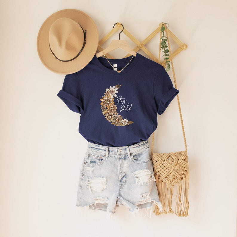 Puede incluir: Una camiseta azul marino con un gr&aacute;fico de luna creciente hecho de flores y el texto "Stay Wild".