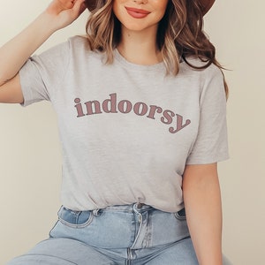 May include: A light gray t-shirt with the word "indoorsy" printed in a mauve color. The shirt is made of a soft material and is paired with blue jeans. The overall style is casual and comfortable.