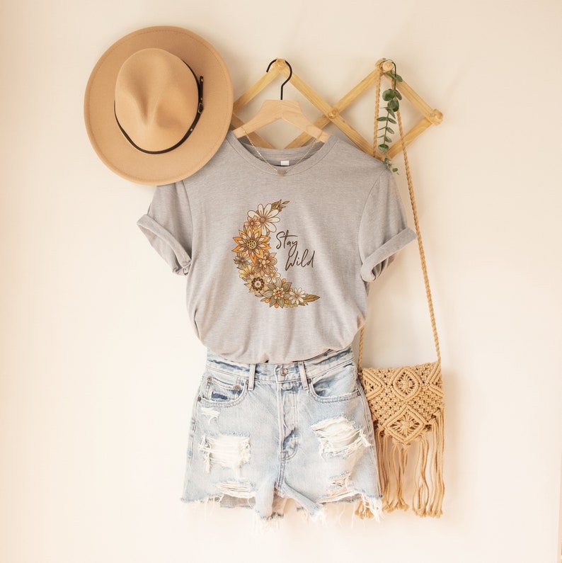 Puede incluir: Una camiseta gris con un dise&ntilde;o de luna floral y el texto "Stay Wild". La camiseta est&aacute; colgada en un estante de madera con un sombrero de paja y una bolsa tejida.