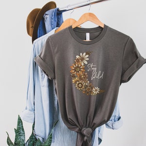 Puede incluir: Una camiseta gris con un dise&ntilde;o de luna creciente hecha de flores naranja, amarilla y marr&oacute;n. El texto "Stay Wild" est&aacute; escrito en blanco en la luna.