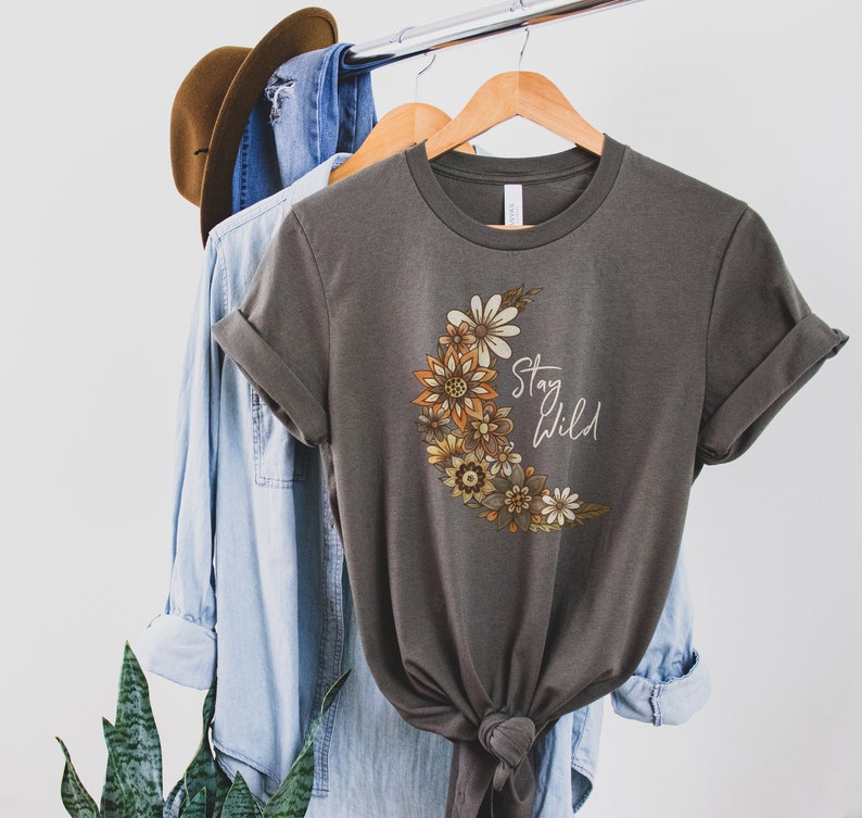 Puede incluir: Una camiseta gris con un dise&ntilde;o de luna creciente hecha de flores naranja, amarilla y marr&oacute;n. El texto "Stay Wild" est&aacute; escrito en blanco en la luna.