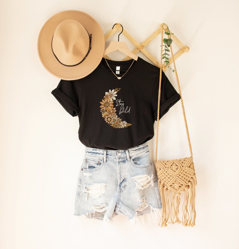 Puede incluir: Camiseta negra con un gr&aacute;fico de luna creciente hecho de flores y el texto "Stay Wild". La camiseta est&aacute; colgada en una percha de madera con un sombrero de paja y una bolsa tejida.