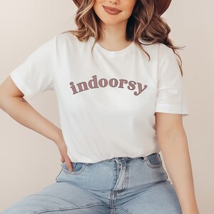 May include: White t-shirt with the word "indoorsy" printed in a mauve color. The shirt has short sleeves and a crew neck. The text is in a rounded, sans-serif font.