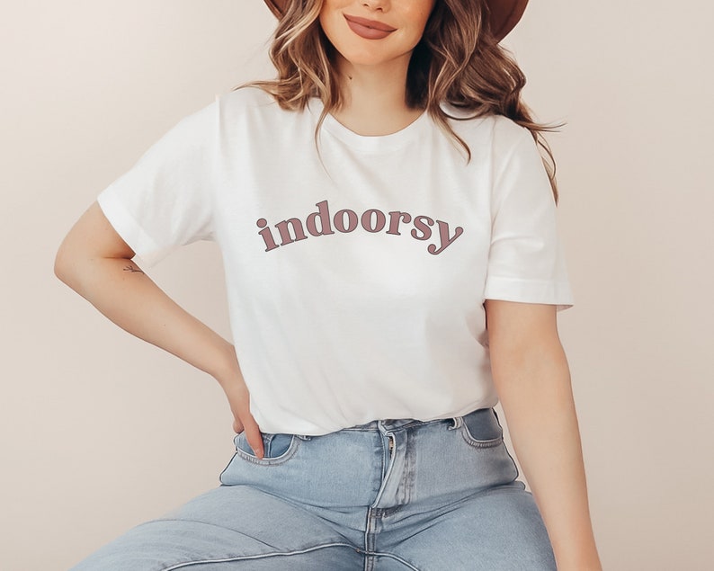 May include: White t-shirt with the word "indoorsy" printed in a mauve color. The shirt has short sleeves and a crew neck. The text is in a rounded, sans-serif font.