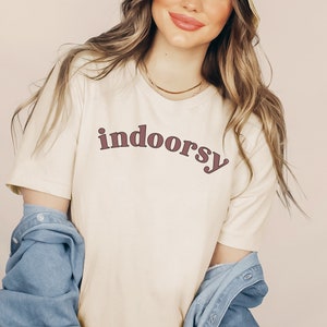 May include: Cream-colored t-shirt with the word "indoorsy" in a burgundy retro font. The shirt has short sleeves and is paired with a denim jacket. A gold necklace is visible.
