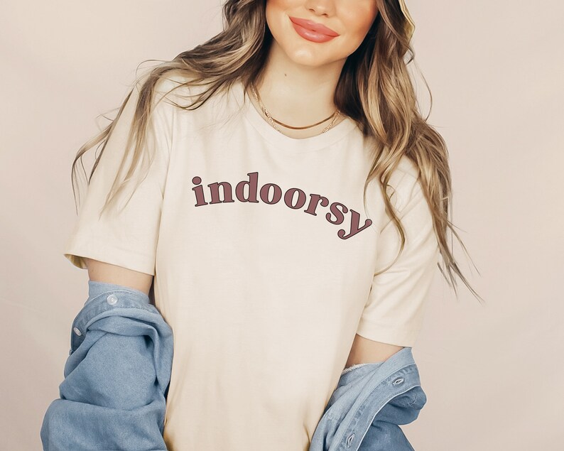 May include: Cream-colored t-shirt with the word "indoorsy" in a burgundy retro font. The shirt has short sleeves and is paired with a denim jacket. A gold necklace is visible.