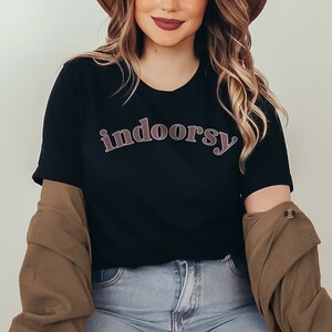 May include: Black t-shirt with the word "indoorsy" in a curved, retro-style font. The text is in a reddish-brown color with a lighter outline. The shirt is paired with a brown jacket and blue jeans.
