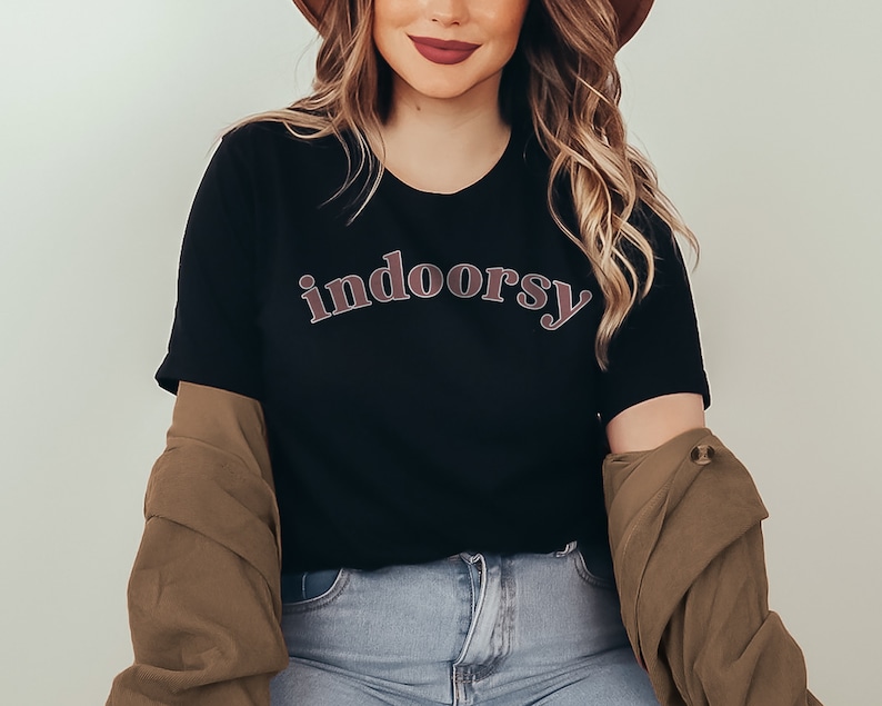 May include: Black t-shirt with the word "indoorsy" in a curved, retro-style font. The text is in a reddish-brown color with a lighter outline. The shirt is paired with a brown jacket and blue jeans.
