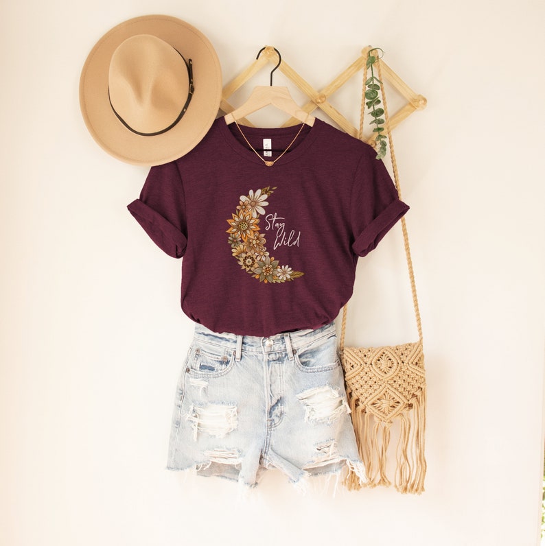 Puede incluir: Una camiseta color burdeos con un gr&aacute;fico de luna creciente con detalles florales en tonos marrones, amarillos y blancos. El texto "Stay Wild" est&aacute; impreso en blanco en la camiseta.