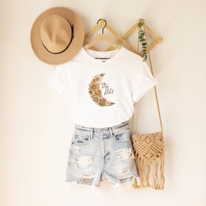 Puede incluir: Camiseta blanca con un gr&aacute;fico de luna creciente hecho de flores y el texto "Stay Wild".