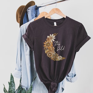 Puede incluir: Una camiseta gris oscuro con un dise&ntilde;o de luna creciente hecho de flores amarillas, naranjas y marrones. El texto "Stay Wild" est&aacute; impreso en blanco en la camiseta.