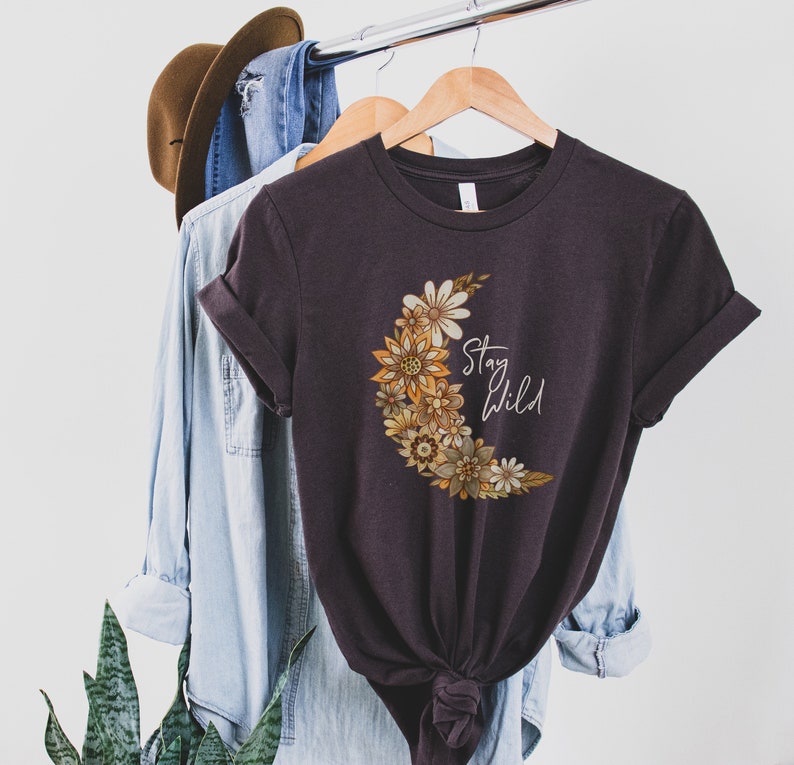 Puede incluir: Una camiseta gris oscuro con un dise&ntilde;o de luna creciente hecho de flores amarillas, naranjas y marrones. El texto "Stay Wild" est&aacute; impreso en blanco en la camiseta.