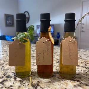 Puede incluir: Tres pequeñas botellas de vidrio con tapas negras y etiquetas de papel marrón. Las botellas contienen líquidos transparentes y están etiquetadas como "Menta", "Azúcar moreno" y "Lavanda".