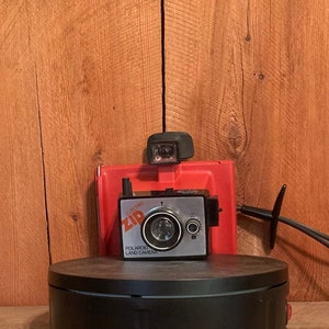 May include: A vintage Polaroid Land Camera with a red case and a black turntable on a wooden shelf.