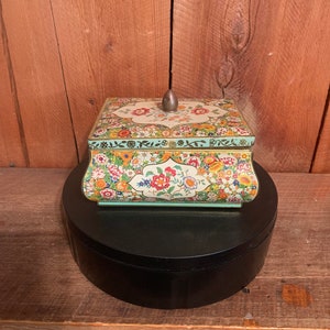 May include: A vintage floral patterned tin box with a raised lid and a small knob on top. The box is sitting on a black circular stand.