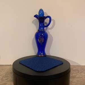 May include: A cobalt blue glass bottle with a gold design and a stopper. The bottle is sitting on a black circular platform with a blue square mat.