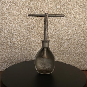 May include: A silver metal nutcracker with a T-shaped handle and a screw mechanism. The nutcracker is on a black surface.