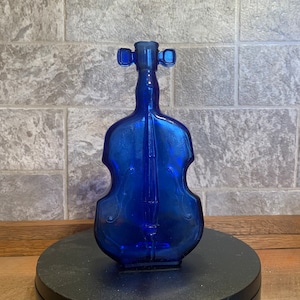 May include: A cobalt blue glass bottle shaped like a violin. The bottle has a clear glass stopper with two small knobs on the top.