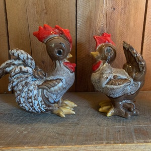 May include: Two ceramic rooster figurines, one brown with white speckles and a red comb, the other brown with white speckles and a red comb, facing each other on a wooden surface.