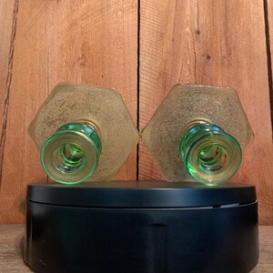 May include: A pair of green glass candle holders with a gold etched design. The candle holders are on a black circular base.