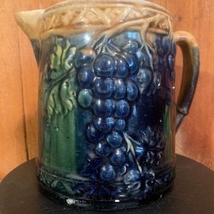 May include: A vintage ceramic pitcher with a blue and green glaze. The pitcher features a raised design of grapes and vines.