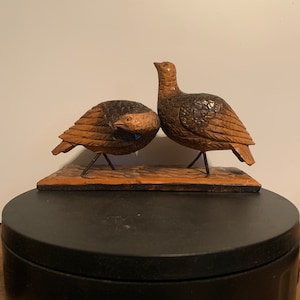 May include: Two intricately carved wooden birds, one brown and one dark brown, perched on a wooden base. The birds are facing different directions, with one looking up and the other looking down.