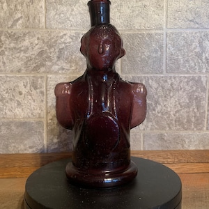 May include: A purple glass bottle shaped like a person with a round head and arms outstretched. The bottle has a small opening at the top.