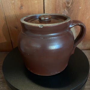 May include: A brown ceramic crock with a lid and handle. The crock has a rounded body and a slightly lighter rim. The lid has a small knob. The crock is sitting on a black surface against a wooden background.