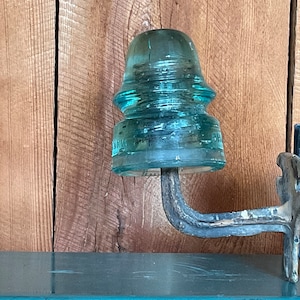 May include: A vintage glass insulator with a light blue tint, mounted on a metal bracket. The insulator has a rounded top and a textured surface. The bracket is made of metal and has a dark gray finish.