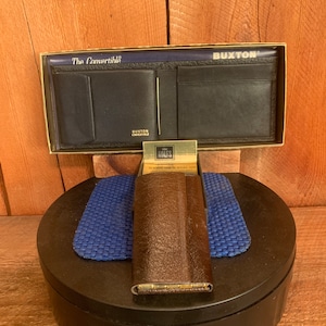May include: A brown leather wallet with a gold metal trim and the words "The Convertible" and "Buxton" printed on the front. The wallet is in a box with a blue and black patterned lining. The box also contains a small gold metal card holder with the word "Rolex" printed on the front.