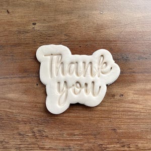 May include: A light-colored cookie cutter in the shape of a cloud, with the words "Thank you" cut out in a cursive font. The cookie cutter is set against a brown wooden background.