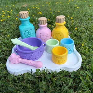 May include: A set of colorful silicone potion-making toys including a purple cauldron, a light pink wand, a light green spoon, three small cups in yellow, green, and blue, and three bottles in teal, pink, and yellow, all on a white tray.